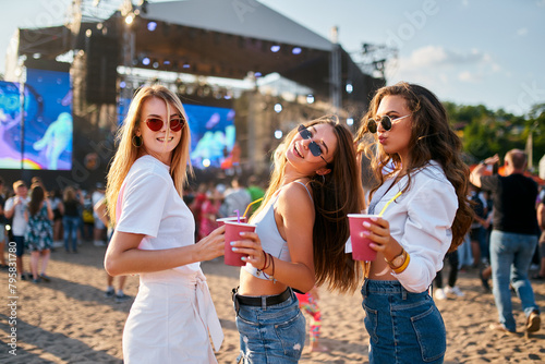Group of smiling young women with sunglasses enjoy summer music festival on beach holding colorful drinks, casual fashion, sunset party vibe with concert stage in background and crowd gathering.