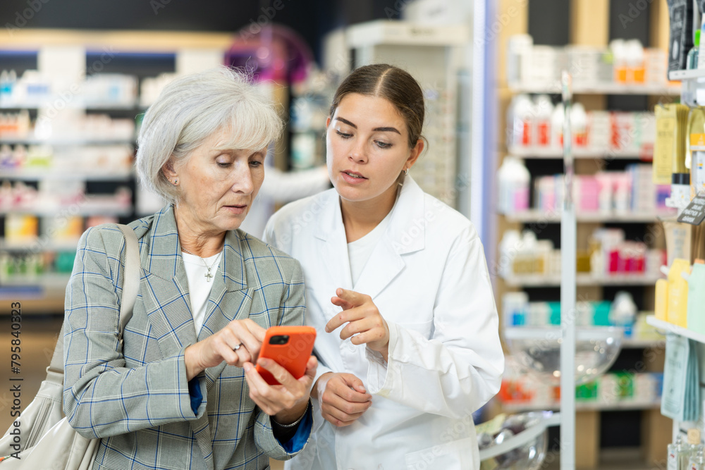 Female pharmacist and elderly woman customer reading prescription together on smartphone screen in pharmacy