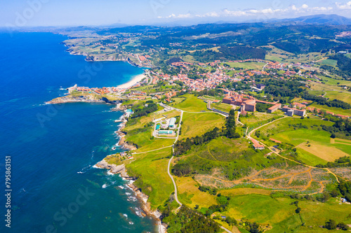 Picturesque aerial view of green valleys on Cantabrian Sea coastal area with brownish roofs of houses of Comillas village, Spain..