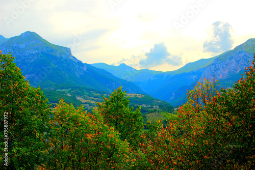 Magical scenery from Montefortino with golden hour cloudy sky in suffused dramatic yellow light, grey clouds, imposing rocky Sibillini mountains and crests, a barrier of dense trees in the foreground