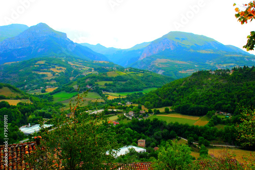 Natural scenery from Montefortino with green trees and rooftops, various white buildings, hills topped by dense thickets alternating with fields and meadows, and the spectacular Sibillini mountains