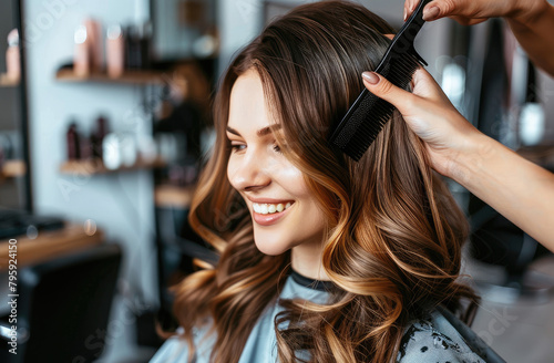 A woman is getting her hair styled at the salon, smiling and looking happy as she sits in an armchair while another person combs their long wavy blonde hair with a black comb