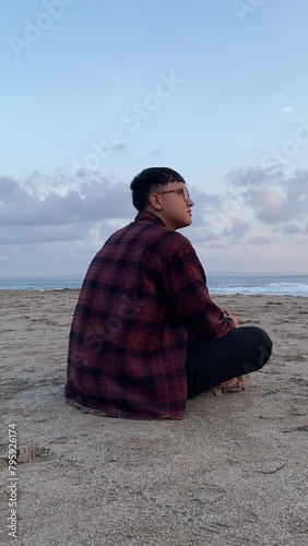 Young person sitting contemplatively on sandy beach looking out at calm ocean water during golden hour with a cloudy sky