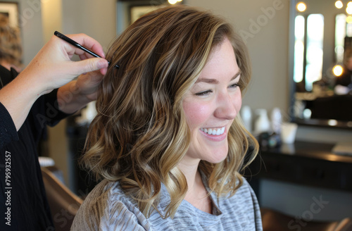 A woman is getting her hair styled at the salon, smiling and looking happy as she sits in an armchair while another person combs their long wavy blonde hair with a black comb