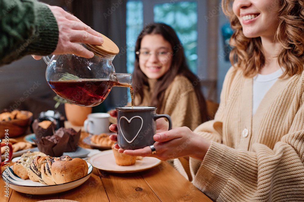 Hand of unrecognizable man holding glass teapot pouring tea into mug during family dinner at home