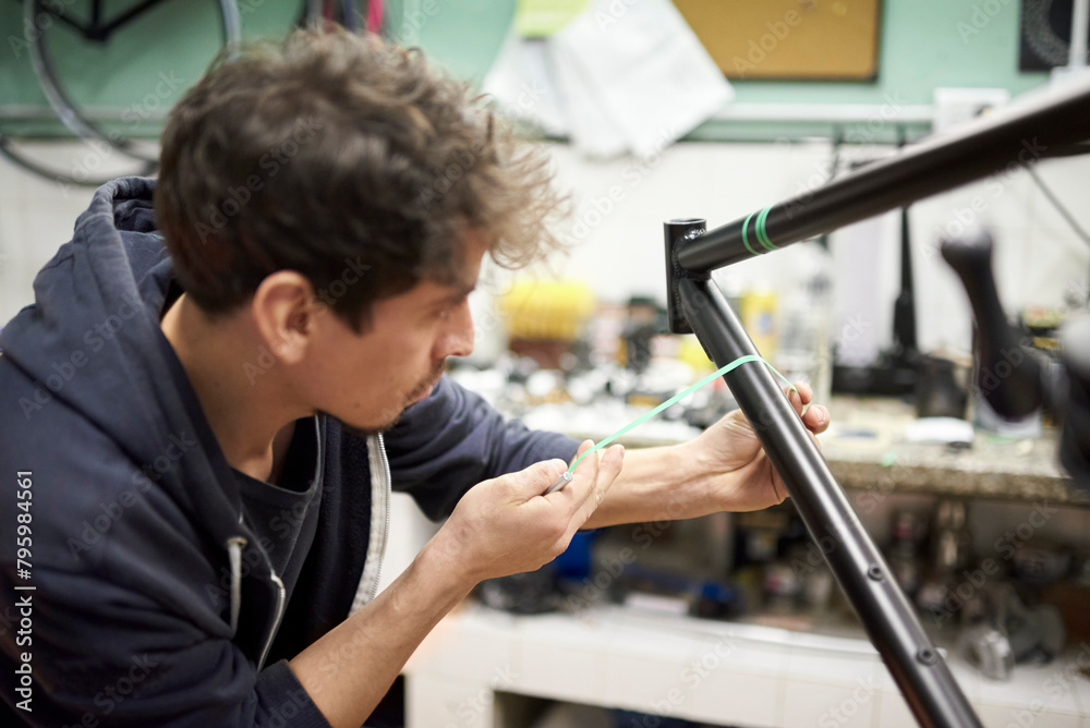 Hispanic young man preparing a bicycle frame with masking tape for a custom painting design in his bike workshop, a handcrafted creative process. Hispanic young man preparing a bicycle frame with masking tape for a custom painting design in his bike workshop, a handcrafted creative process.
