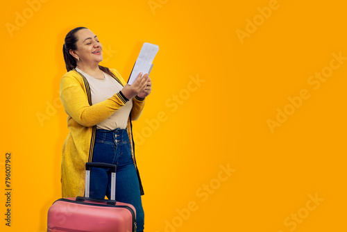 Beautiful woman in summer clothes with pink suitcase, happy with her hand up, celebrating her cheap ticket on yellow background. Tourist very happy to travel abroad with a bargain ticket. Copy space. photo