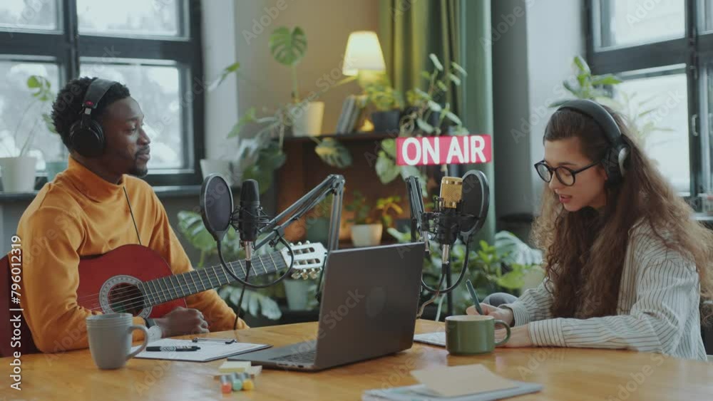 Young Caucasian female podcaster in headphones talking in mic and interviewing Black musician sitting with guitar in recording studio Young Caucasian female podcaster in headphones talking in mic and interviewing Black musician sitting with guitar in recording studio