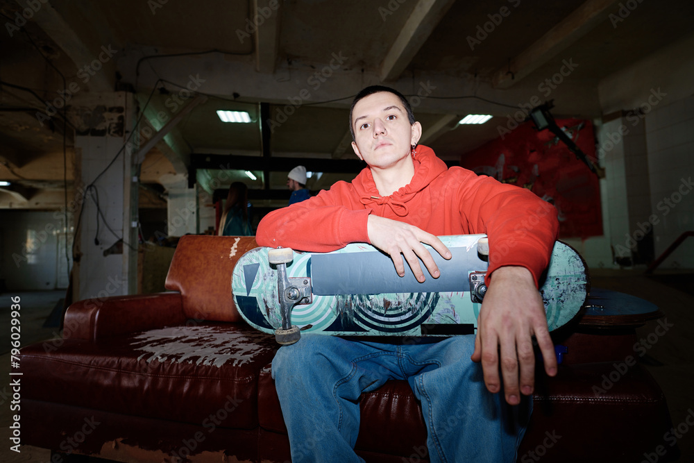 Obraz premium Flash photo of self-confident gen Z man wearing red hoodie and jeans sitting on shabby couch in skatepark looking at camera