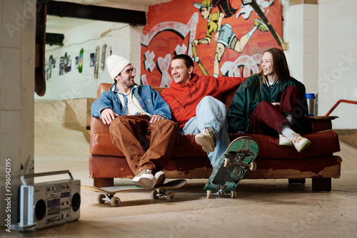 Group of three modern male and female friends hanging out in skatepark sitting on couch, chatting and listening to music, copy space