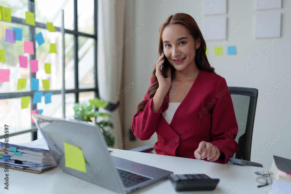 Happy young asian woman talking on the mobile phone and smiling while sitting at her working place in office. Happy young asian woman talking on the mobile phone and smiling while sitting at her working place in office.