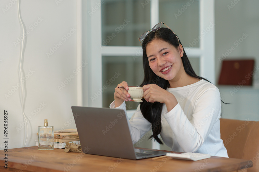 Attractive businesswoman holding a glass of hot drink and working on a laptop, relaxing, smiling while sitting in the office in the morning working lifestyle concept. Attractive businesswoman holding a glass of hot drink and working on a laptop, relaxing, smiling while sitting in the office in the morning working lifestyle concept.