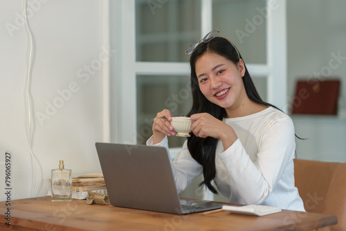 Attractive businesswoman holding a glass of hot drink and working on a laptop, relaxing, smiling while sitting in the office in the morning working lifestyle concept.