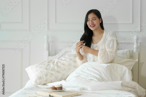 Asian woman wakes up in the morning on a white bed and looks in the mirror to see her beauty. Spray on perfume for freshness. Feel happy in beauty Health care concepts, lifestyle, skin care.