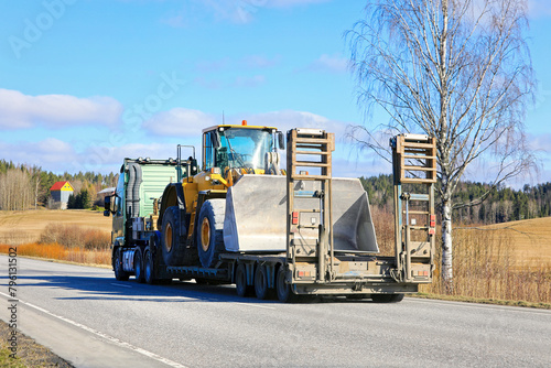 Truck Transports Wheel Loader on Low Loader Semitrailer along Highway on a Day of Spring.