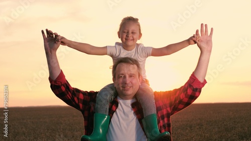 Smiling father and daughter walking at sunset agriculture wheat field closeup. Cute little girl sitting on male parent shoulders with open hands enjoy freedom happy childhood sunrise sky meadow