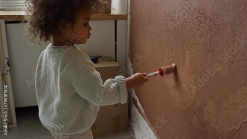 Tracking side footage of little curious Biracial baby girl with curly hair coming up to wall with paintbrush and painting it with brown color during renovation process