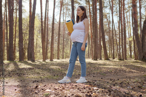 Full length of pregnant woman wearing casual clothing holding yoga mat while standing in spring forest preparing for workout in open air enjoying yoga in wood © sementsova321