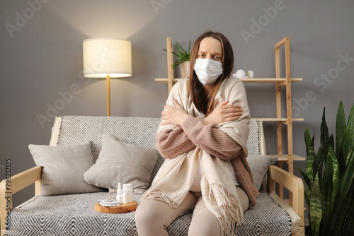 Sick unhealthy pregnant woman wearing medical mask suffering flu symptoms and high temperature sitting on couch wrapped in blanket feeling cold in cozy living room at home