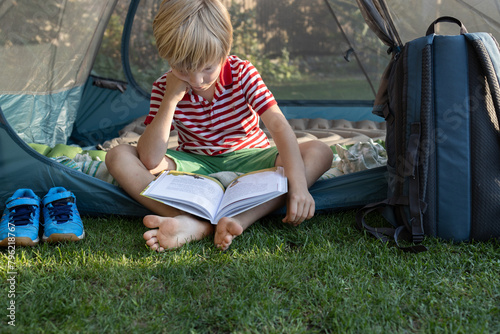 young barefoot child reads a book with interest while sitting in a tent on a warm sunny day. Summer leisure, adventure and outdoor recreation Passion for reading
