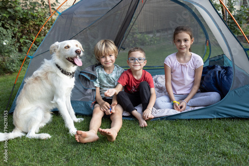 Three happy children and a dog are sitting in a tent on a warm sunny day. Summer active family fun, adventure and outdoor recreation. Children's camping with favorite pet. Healthy lifestyle