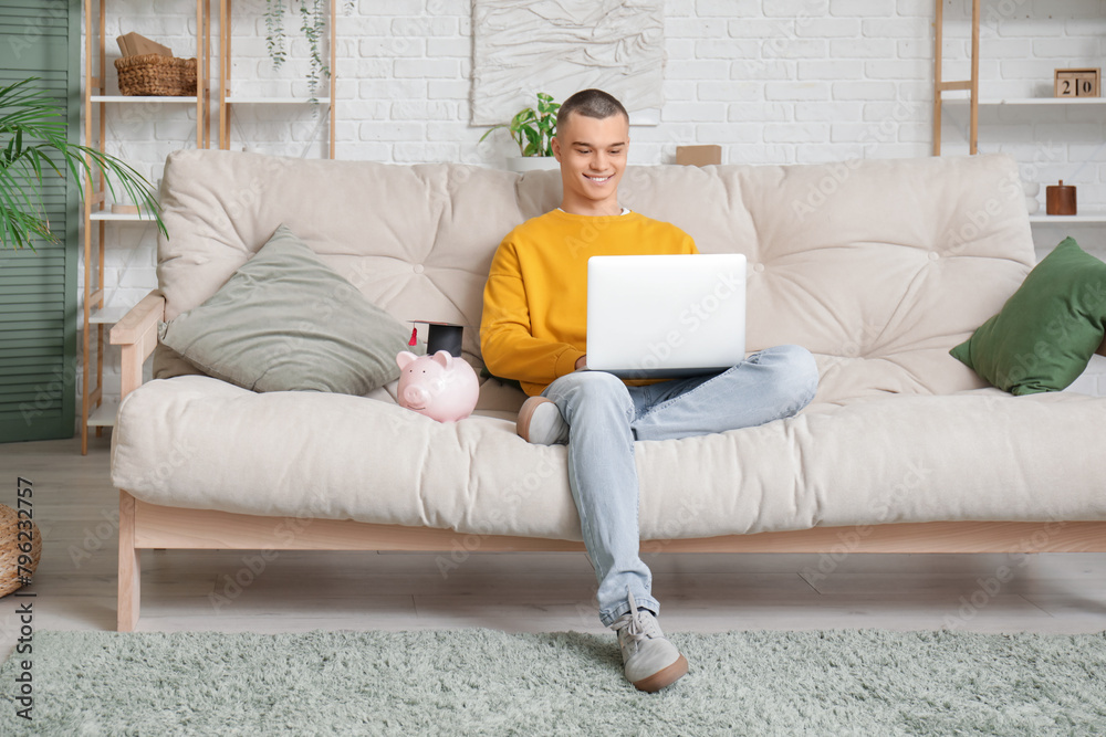 Male student with piggy bank and laptop on sofa in room. Concept of savings for education Male student with piggy bank and laptop on sofa in room. Concept of savings for education