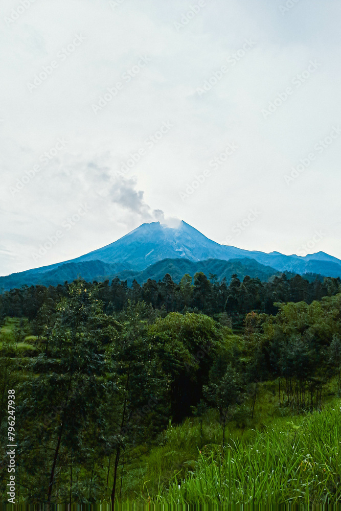 Mount Merapi in Indonesia in the afternoon is very clear and majestic against a background of blue sky and green forest with a ravine flowing cold lava in the foreground.