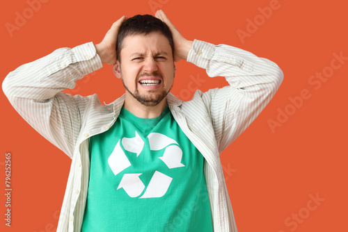 Angry young man in t-shirt with recycling logo holding head on orange background. Ecology concept
