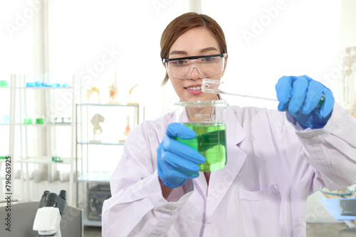 Portrait of happy beautiful Asian scientist woman in lab coats with safety glasses do science experiments. Researcher pour chemical from tube into beaker for research, female work in science field.