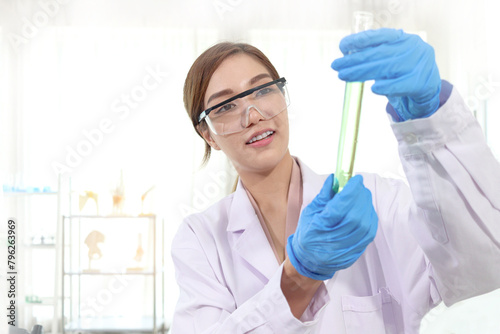 Beautiful Asian scientist woman in lab coats with safety glasses holding test tube with green chemical. Researcher uses equipment to do science experiment for research. Female works in science field.