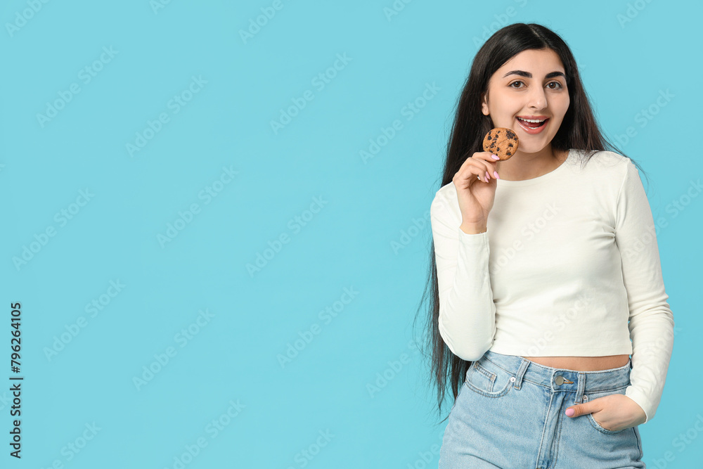 Fototapeta premium Beautiful young happy woman holding tasty cookie with chocolate chips on blue background