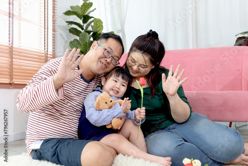 Happy chubby Asian family. Father and mother playing with little girl daughter at home. Parents spending time together with kid, hugging to show love. Child, dad and mom waving hands in living room.
