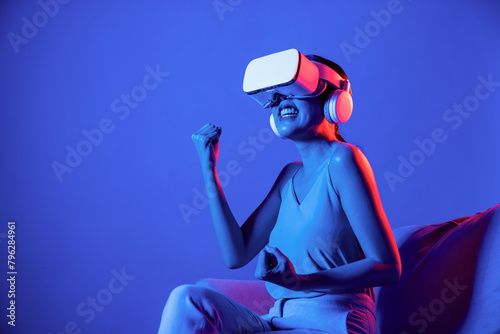 Smart Female sitting on sofa surrounded by neon light wear VR headset connecting metaverse, futuristic cyberspace community technology, watch movie with cheerful action and happy face. Hallucination.