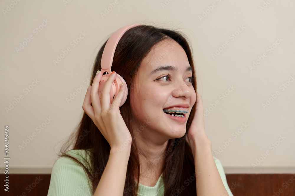 Asian Female in casual attire, touching pink headphone, looking aside, a mix of curiosity and joy on her face. Young listener adjusting her pale headset, glancing away, expressions of wonder Asian Female in casual attire, touching pink headphone, looking aside, a mix of curiosity and joy on her face. Young listener adjusting her pale headset, glancing away, expressions of wonder