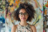 A smiling young woman with curly hair and glasses exudes friendliness amidst an art studio filled with colorful paintings