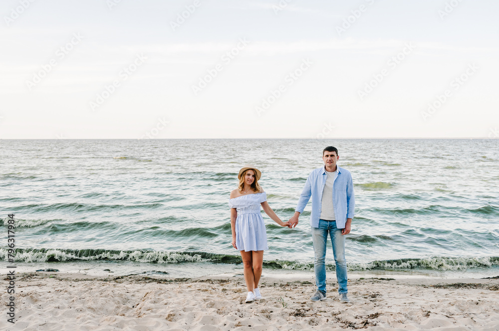 Couple in love holding hands and looking at camera on seashore sea. Man and woman on sand. Female and male standing on beach ocean and enjoying sunny summer day on vacation. Spending time together Couple in love holding hands and looking at camera on seashore sea. Man and woman on sand. Female and male standing on beach ocean and enjoying sunny summer day on vacation. Spending time together