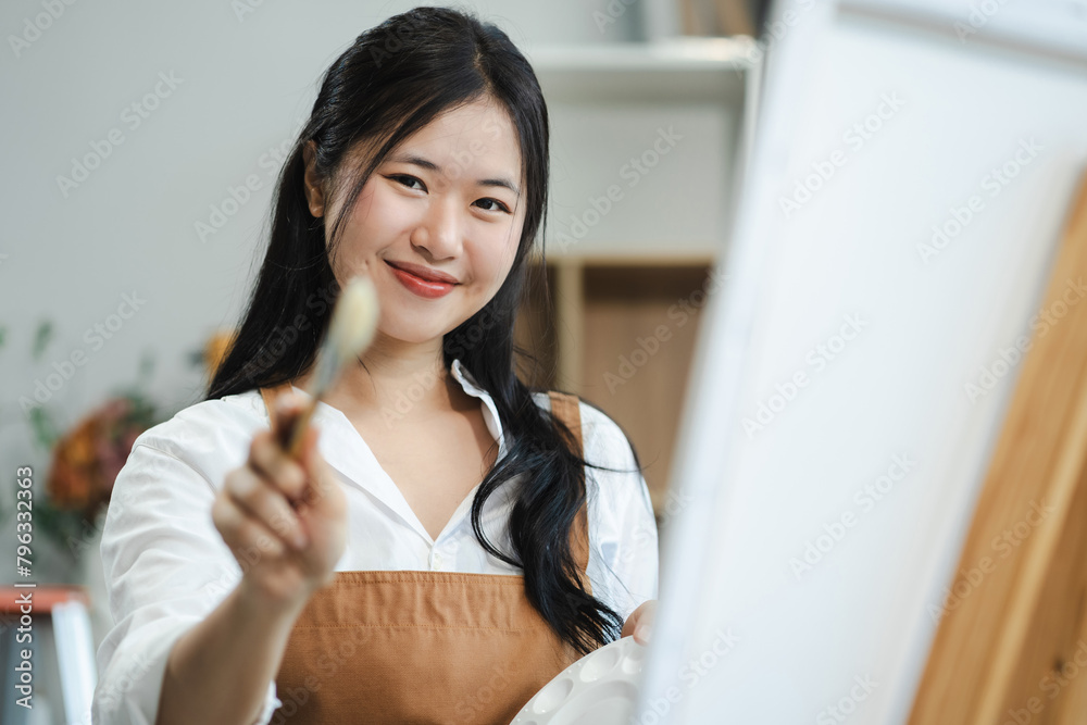 Creative painter painting in an art studio. Artistic young woman working on a new painting. Cheerful young artist looking at the camera while holding a paintbrush. Creative painter painting in an art studio. Artistic young woman working on a new painting. Cheerful young artist looking at the camera while holding a paintbrush.