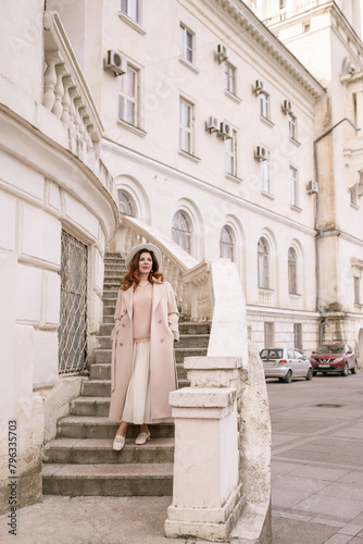 woman in elegant coat and hat against an intricate architectural backdrop, harmoniously blending modern fashion with historical allure. The soft daylight adds to its timeless appeal.