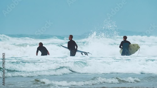 Three surfers walk on the beach with surfboards and enter into the ocean