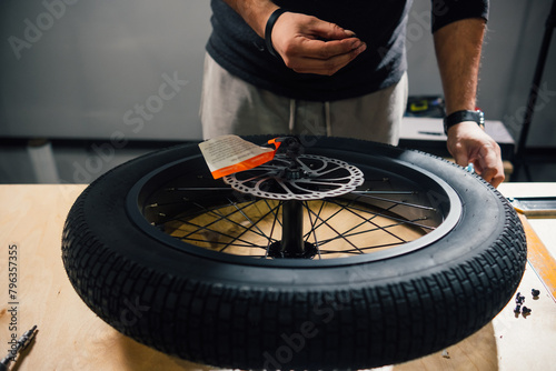 Closeup view of new motorcycle wheel lies on the table in the workshop. The craftsman is holding screw in the hand trying to mount brake systems disk