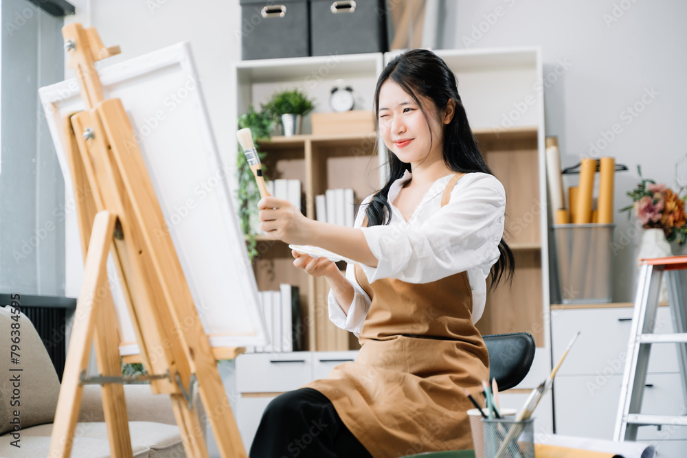 Asian Female painter do artwork in art workshop, painting supplies, oil pastels, two canvas easel, creative space with paintbrush in art studio