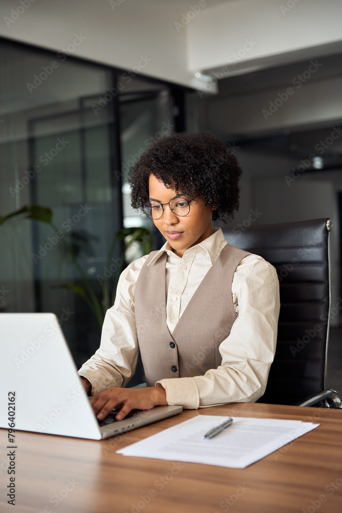 Busy African business woman using laptop working in corporate office. Young businesswoman corporate executive at work, professional female company hr or bank manager looking at computer. Vertical. Busy African business woman using laptop working in corporate office. Young businesswoman corporate executive at work, professional female company hr or bank manager looking at computer. Vertical.