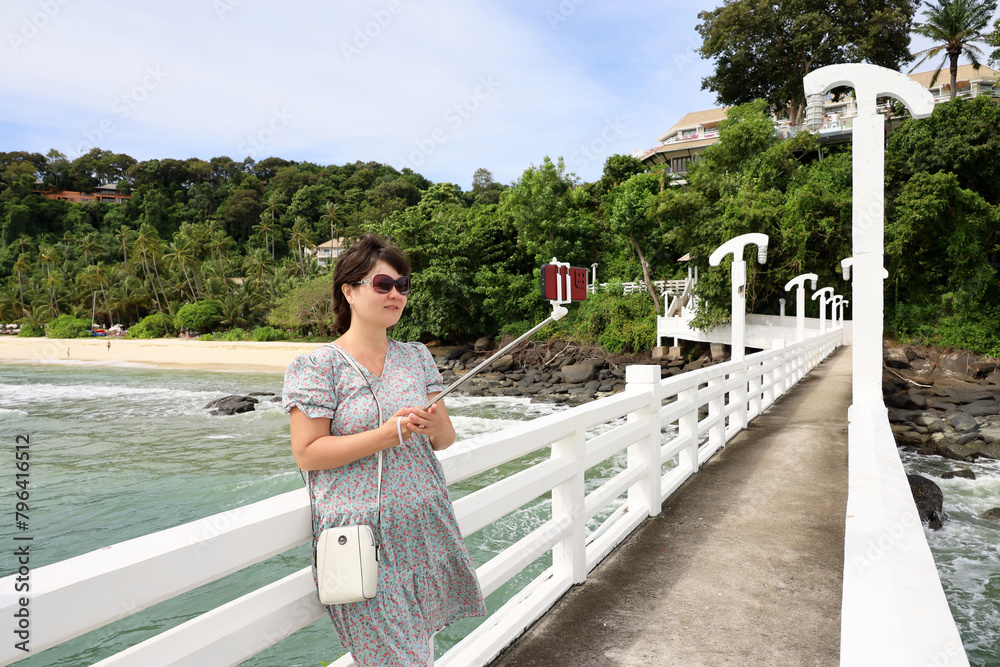 Woman in sunglasses taking selfie on smartphone camera standing on a pier on tropical sea beach