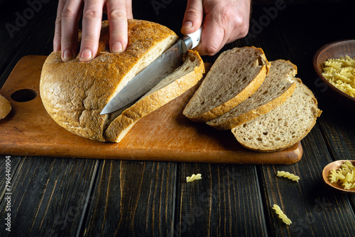 Slicing rye bread on a kitchen board with a knife in the hands of a cook before breakfast.