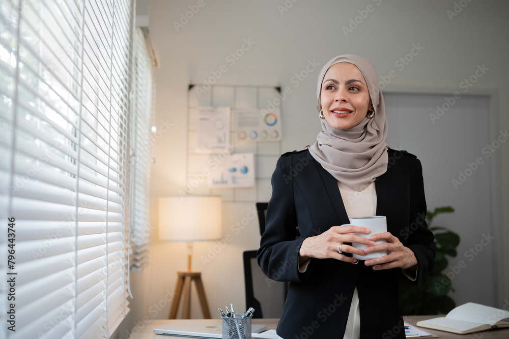 Confident Muslim businesswoman standing with coffee mug in front of desk in office during morning time Confident Muslim businesswoman standing with coffee mug in front of desk in office during morning time