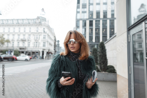 stylish urban beautiful fashionable girl with glasses in a fashion shaggy jacket with a magazine and a smartphone walks in the city