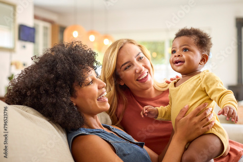 Same Sex Female Couple Or Friends Playing With Baby Sitting On Sofa At Home Together