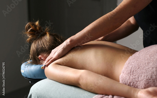 Professional Masseuse Massaging Back And Shoulder Blades Of Young Woman Lying On A Massage Table.