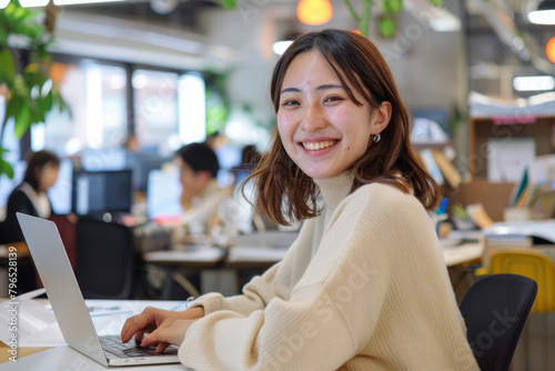 Positive Japanese woman working on a laptop in a cheerful office environment, reflecting success and positivity.