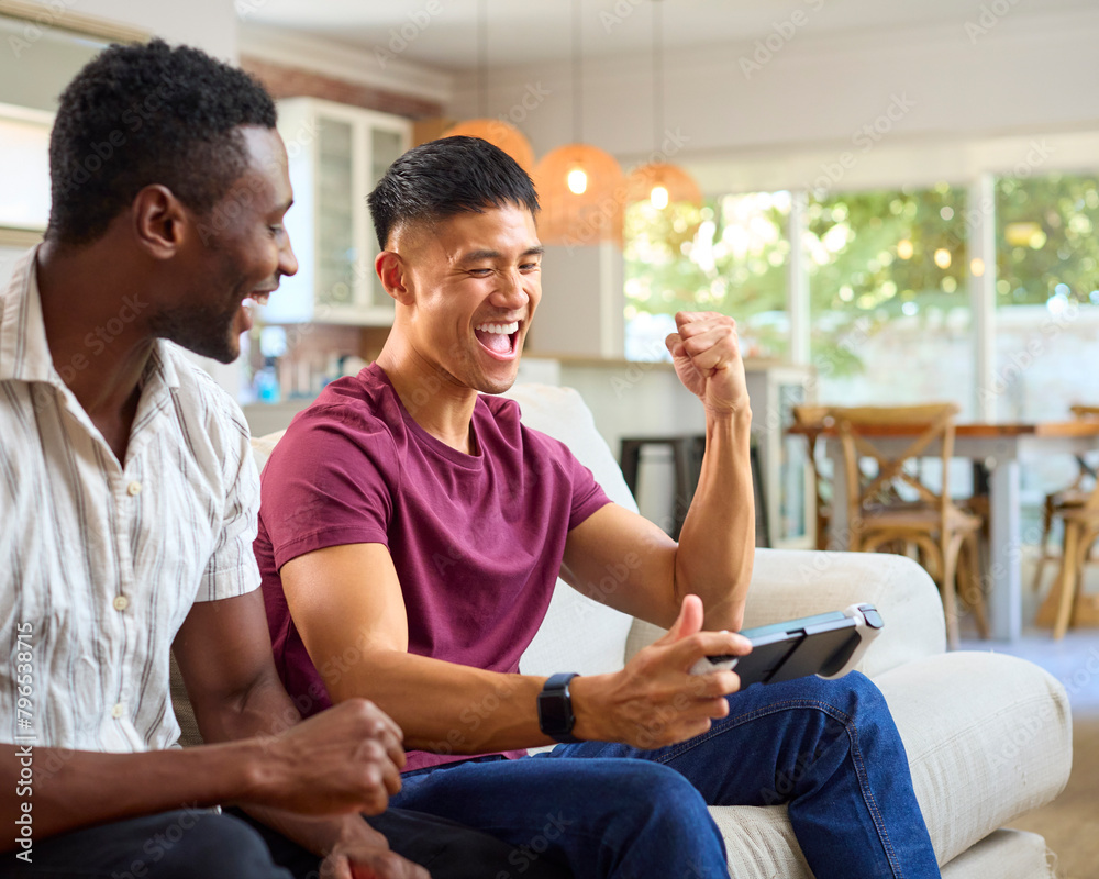 Same Sex Male Couple Or Friends On Sofa At Home With Handheld Mobile Computer Gaming Console